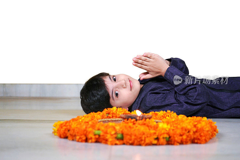 一个快乐微笑的印度亚洲男孩看着一个Aarti Pooja或祈祷塔利与火焰，Jyot, Diya, Deepak火焰和金盏花躺在地板上，做合十礼孤立的白色